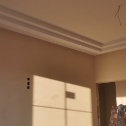 Rafał Jędrzejewski - Wnętrze w trakcie remontu: świeżo pomalowane ściany w beżowym odcieniu z widocznymi otworami na instalacje, ozdobne listwy przysufitowe i cień okna na ścianie, drewniana drabina oparta o ścianę...