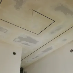 Surowy sufit po nałożeniu gładzi, z widocznymi śladami szpachlowania, obramowany czarnymi listwami, z wystającym przewodem elektrycznym i drabina w rogu pokoju.