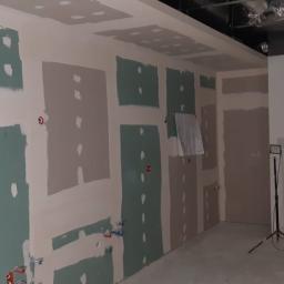 Nf Company Sp. z o.o. - Przygotowanie ścian do dalszych prac wykończeniowych: fragmenty ścian pokryte zielonkawą masą szpachlową, widoczne instalacje elektryczne i hydrauliczne, pracownik na drabinie w tle.