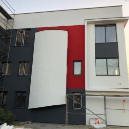 Nf Company Sp. z o.o. - Nowoczesny budynek z częściowo pomalowaną elewacją na biało, szaro i czerwono, widoczne rusztowania i materiały budowlane na ziemi.