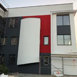 Nowoczesny budynek z częściowo pomalowaną elewacją na biało, szaro i czerwono, widoczne rusztowania i materiały budowlane na ziemi.