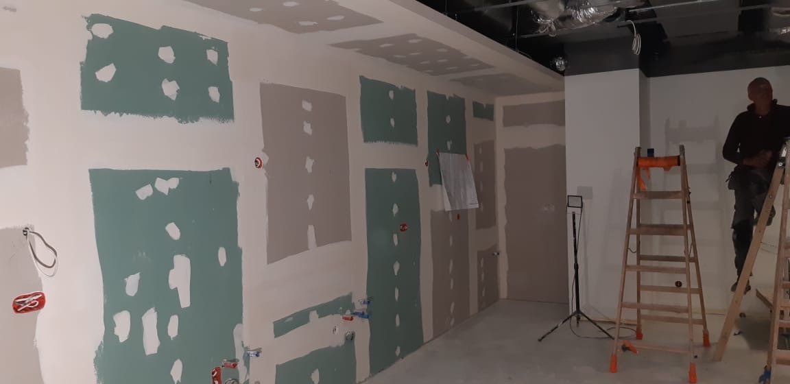 Przygotowanie ścian do dalszych prac wykończeniowych: fragmenty ścian pokryte zielonkawą masą szpachlową, widoczne instalacje elektryczne i hydrauliczne, pracownik na drabinie w tle.