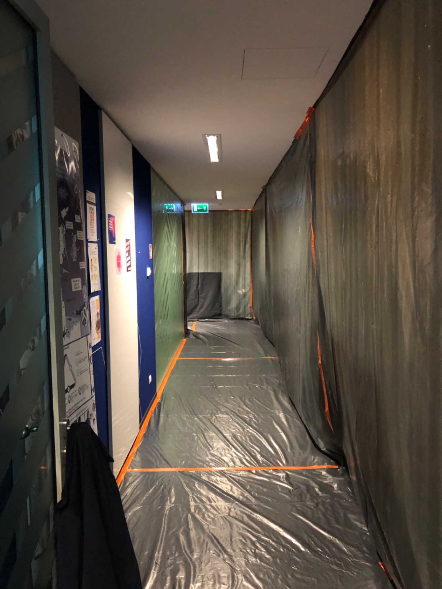 Korytarz biurowy zabezpieczony folią i taśmą malarską przed pracami remontowymi, widoczne plakaty na ścianie i ubranie wiszące na drzwiach.