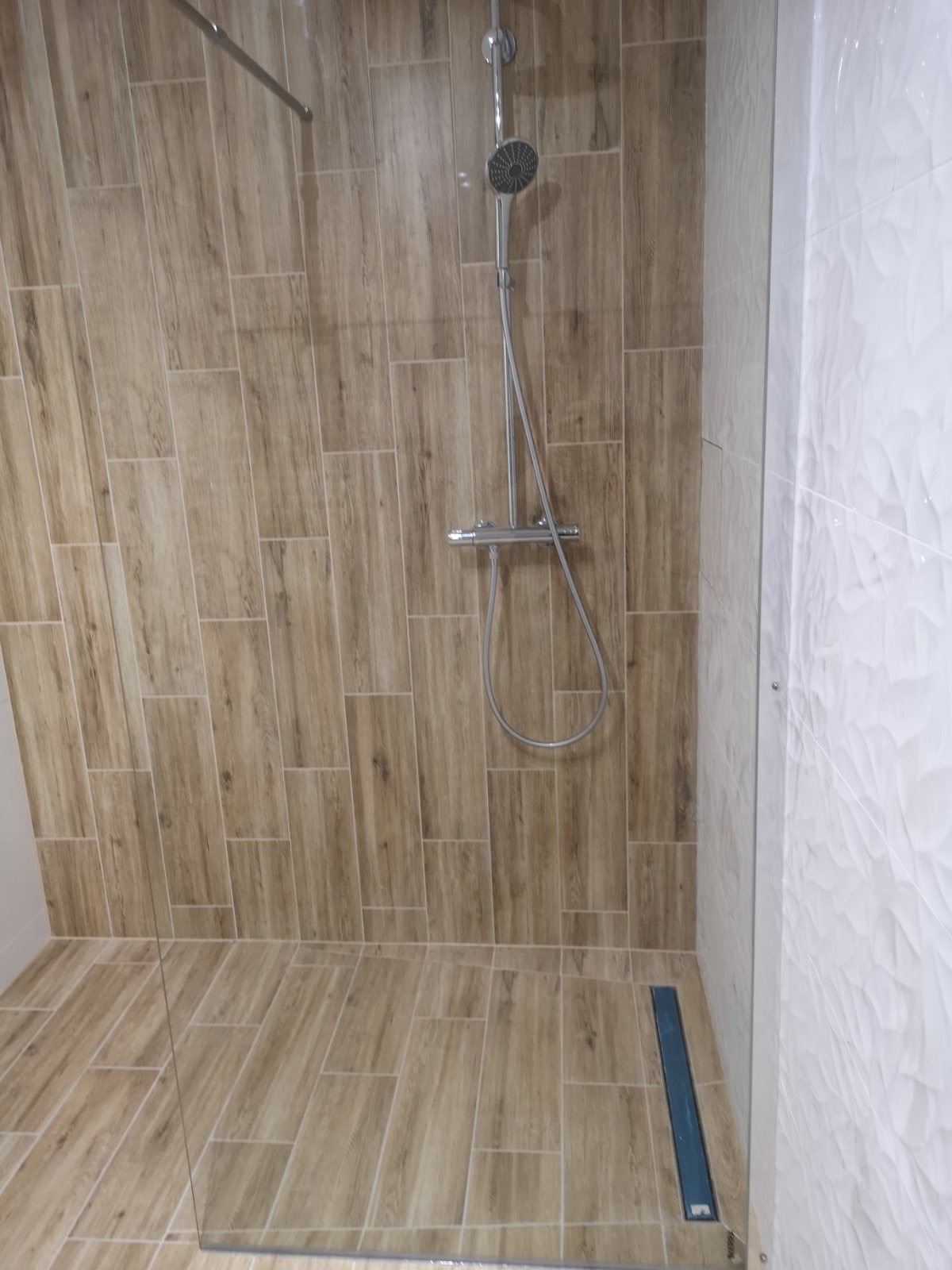 Nowoczesna kabina prysznicowa z drewnopodobnymi płytkami, chromowaną armaturą i odpływem liniowym, wykończona białymi płytkami o falistej fakturze.