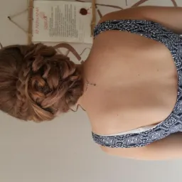 Upięcie włosów z warkoczem na brązowych włosach, widok z tyłu, delikatny naszyjnik, wzorzysta sukienka, na ścianie dekoracja i tekst.