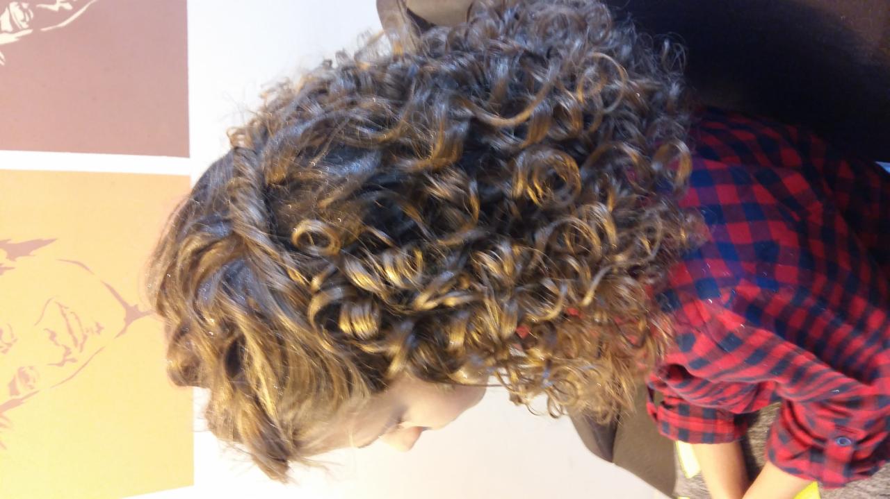 Kobieta z burzą drobnych, błyszczących loków, ubrana w czerwoną koszulę w kratę, pochylona nad czymś poza kadrem, z widocznym fragmentem grafiki na ścianie w tle.