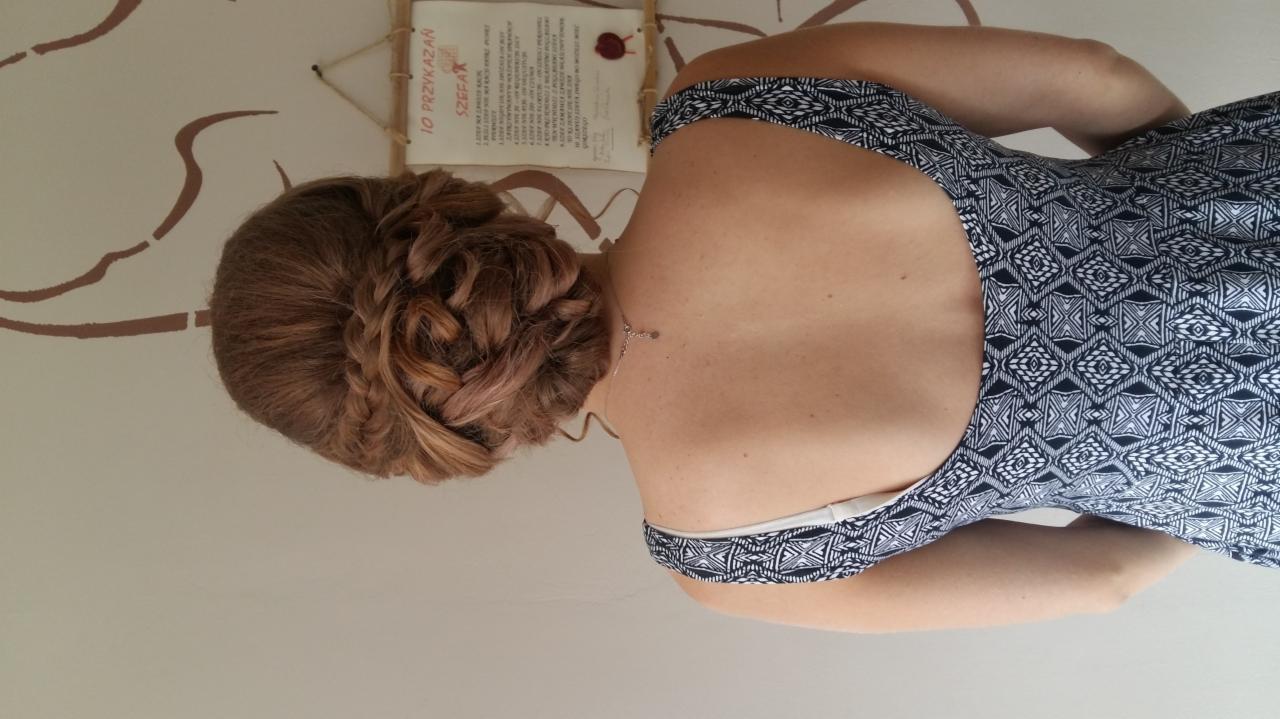 Upięcie włosów z warkoczem na brązowych włosach, widok z tyłu, delikatny naszyjnik, wzorzysta sukienka, na ścianie dekoracja i tekst.