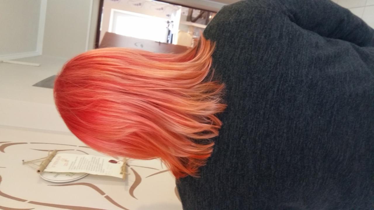 Głowa pochylonej osoby z włosami w odcieniach czerwieni i pomarańczy, widoczne pasemka i tekstura, ciemny sweter, w tle białe ściany i niewyraźny fragment pomieszczenia.