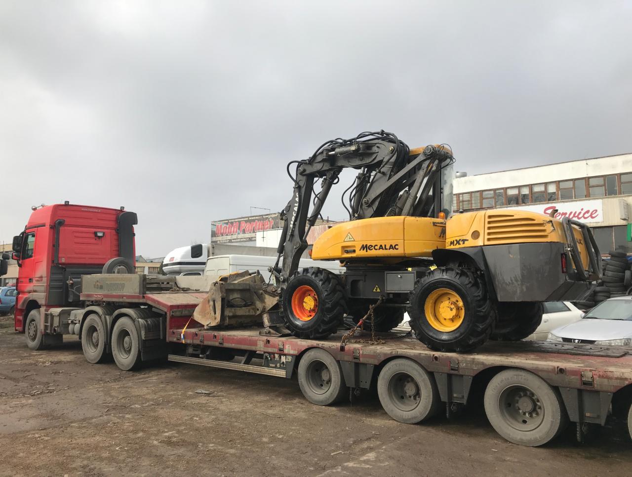 Żółta koparka Mecalac MXT zabezpieczona łańcuchami na niskopodwoziowej platformie transportowej ciągniętej przez czerwony ciągnik siodłowy, widok z boku, pochmurne niebo w tle.