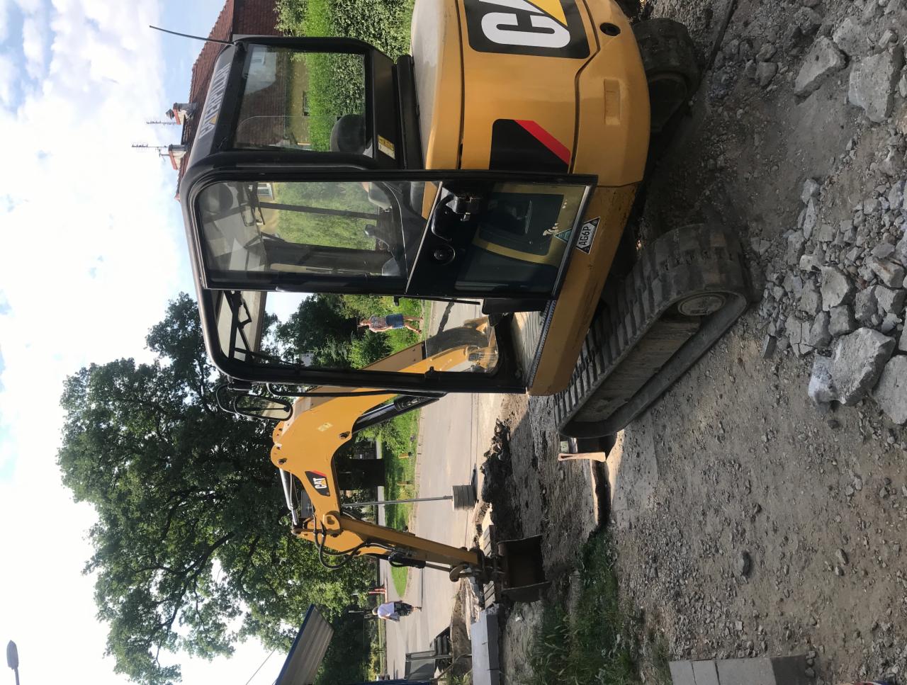 Żółta mini koparka Caterpillar na placu budowy, widoczne ślady prac ziemnych i kostka brukowa w tle, częściowo pochmurne niebo.