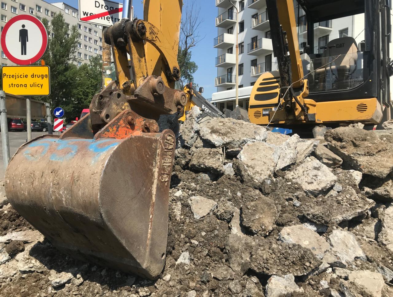 Żółta koparka CAT z brudną łyżką nabiera gruz i ziemię na tle budynków miejskich i znaku drogowego.