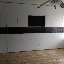 Duża, biała szafa z ciemnobrązowym pasem dekoracyjnym i miejscem na telewizor, wbudowana w ścianę pokoju z parkietem w geometryczny wzór.