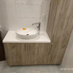 Nowoczesna łazienka z okrągłą umywalką nablatową na białym blacie i szafką z drewnopodobnym fornirem, minimalistyczne białe płytki na ścianie.