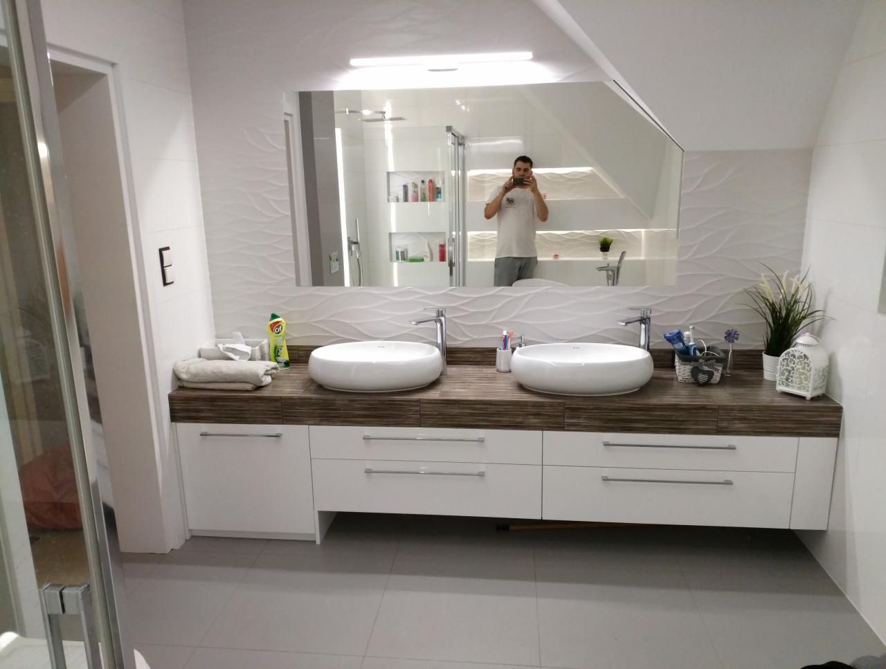 Nowoczesna łazienka z podwójną umywalką nablatową na drewnianym blacie, białe szafki z poziomymi uchwytami, lustro z odbiciem osoby robiącej zdjęcie, dekoracyjne białe płytki ścienne z falistym...