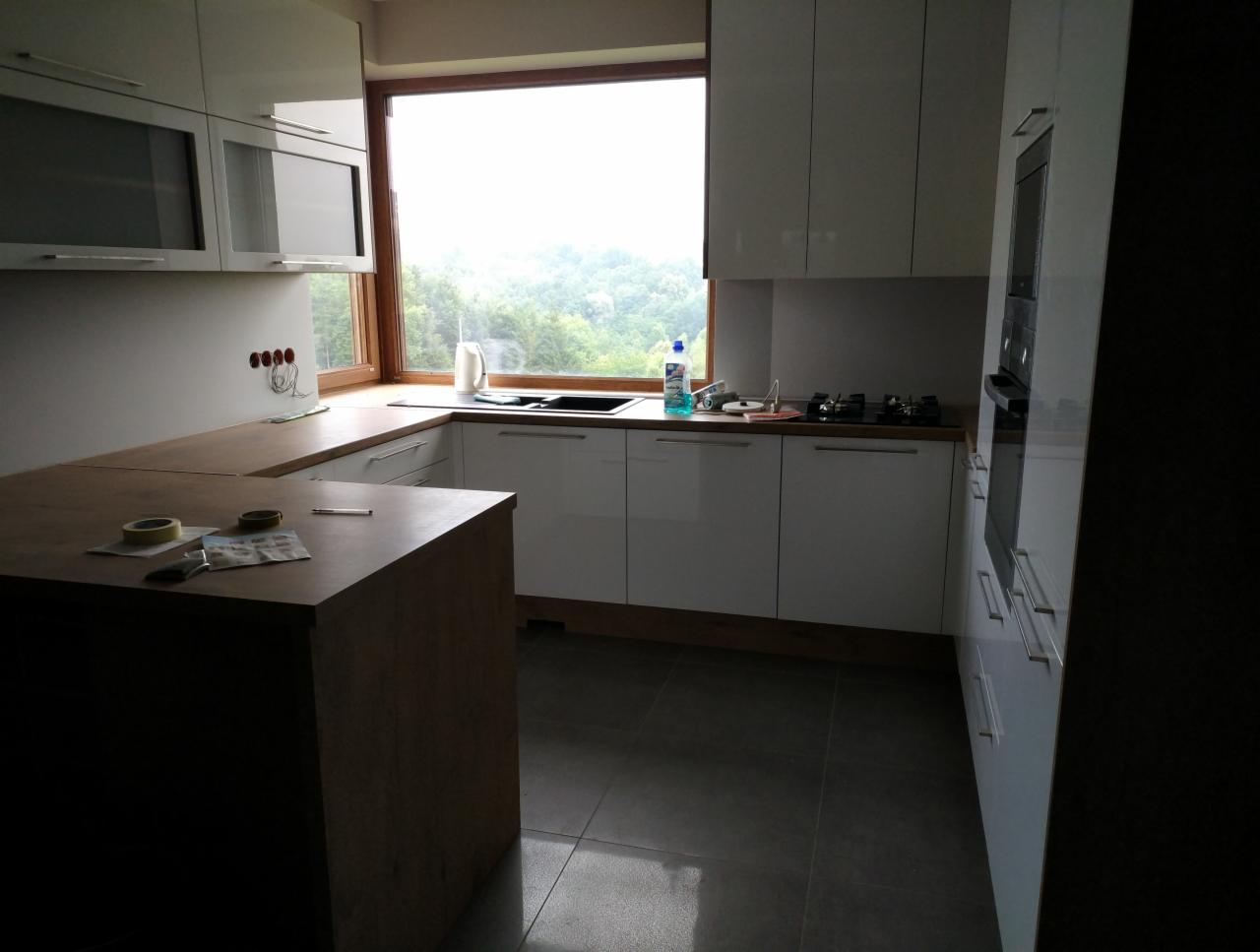 Nowoczesna kuchnia z białymi, lakierowanymi frontami, drewnianym blatem i wyspą, widok na zewnątrz przez duże okno, w tle zieleń. Na wyspie taśma malarska i narzędzia.