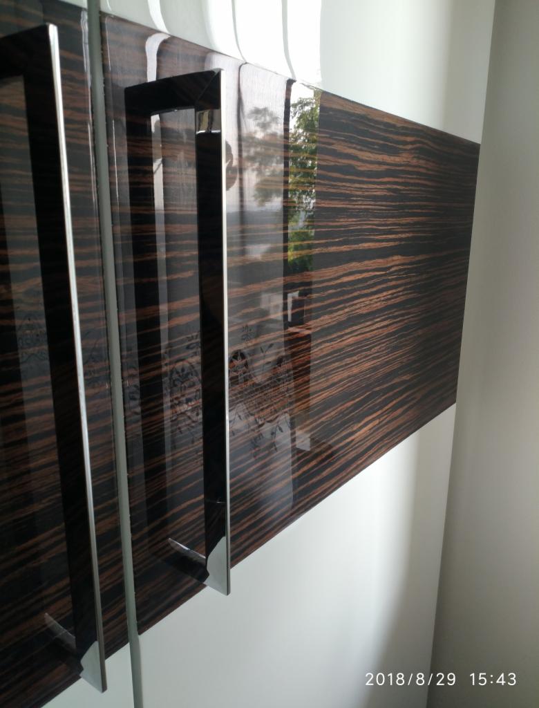 Fragment szafy z fornirem o wzorze zebry, z długimi, prostokątnymi uchwytami ze stali nierdzewnej i subtelnym wzorem kwiatowym na powierzchni.