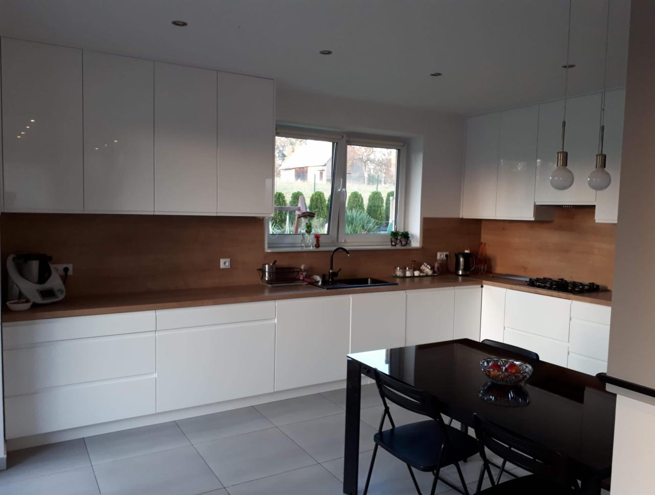 Nowoczesna kuchnia z białymi, lakierowanymi frontami szafek i drewnianym blatem, uzupełniona czarnym stołem z owocami w misie oraz widokiem na ogród przez okno.