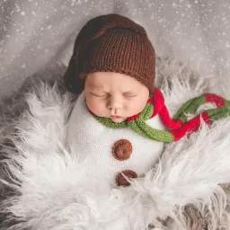 Śpiący noworodek w brązowej czapce, owinięty w biały materiał z guzikami, stylizowany na bałwana, z zielono-czerwonym szalikiem, na tle imitującym śnieg.