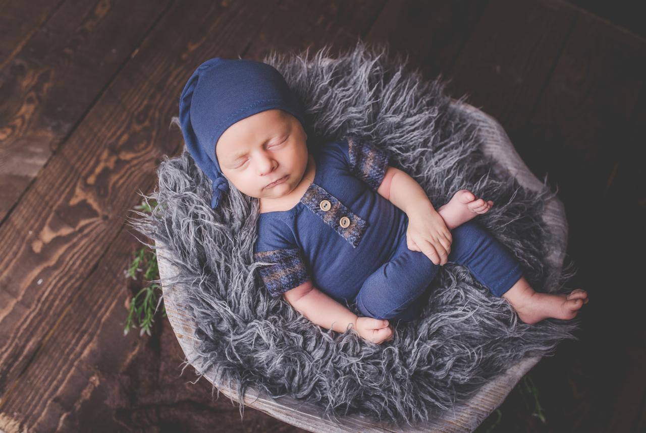 Śpiący noworodek w niebieskim ubranku i czapeczce, ułożony w drewnianej misie wyłożonej szarą, futrzaną tkaniną, na tle drewnianej podłogi.