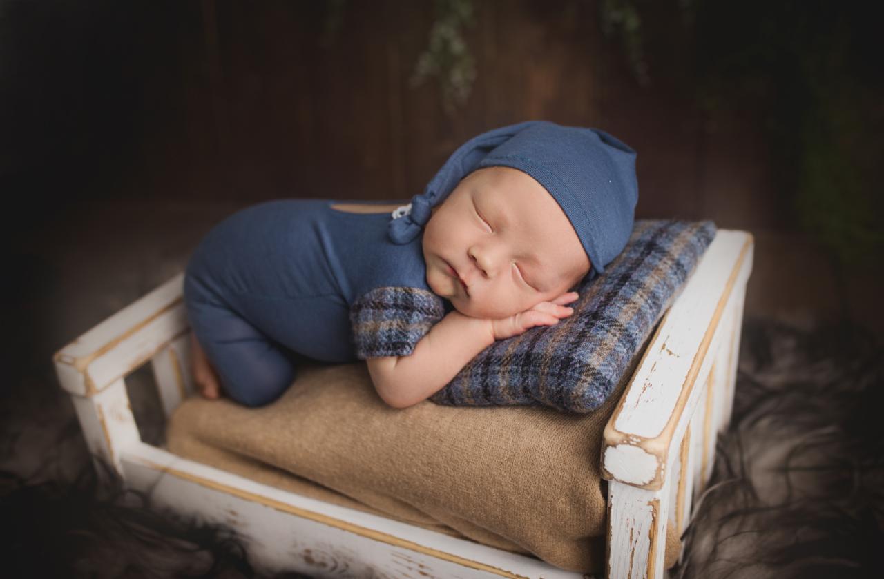 Śpiący noworodek w niebieskim ubranku i czapeczce, leżący na boku na poduszce w małym, białym, drewnianym łóżeczku z dekoracyjnym tłem.
