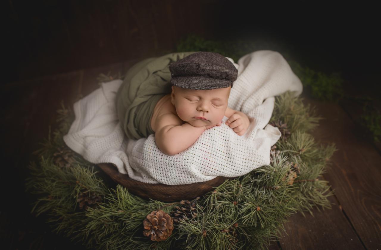 Śpiący noworodek w brązowej czapce leży w drewnianej misce na białym kocyku, otoczony gałązkami sosny i szyszkami na ciemnym drewnianym tle.