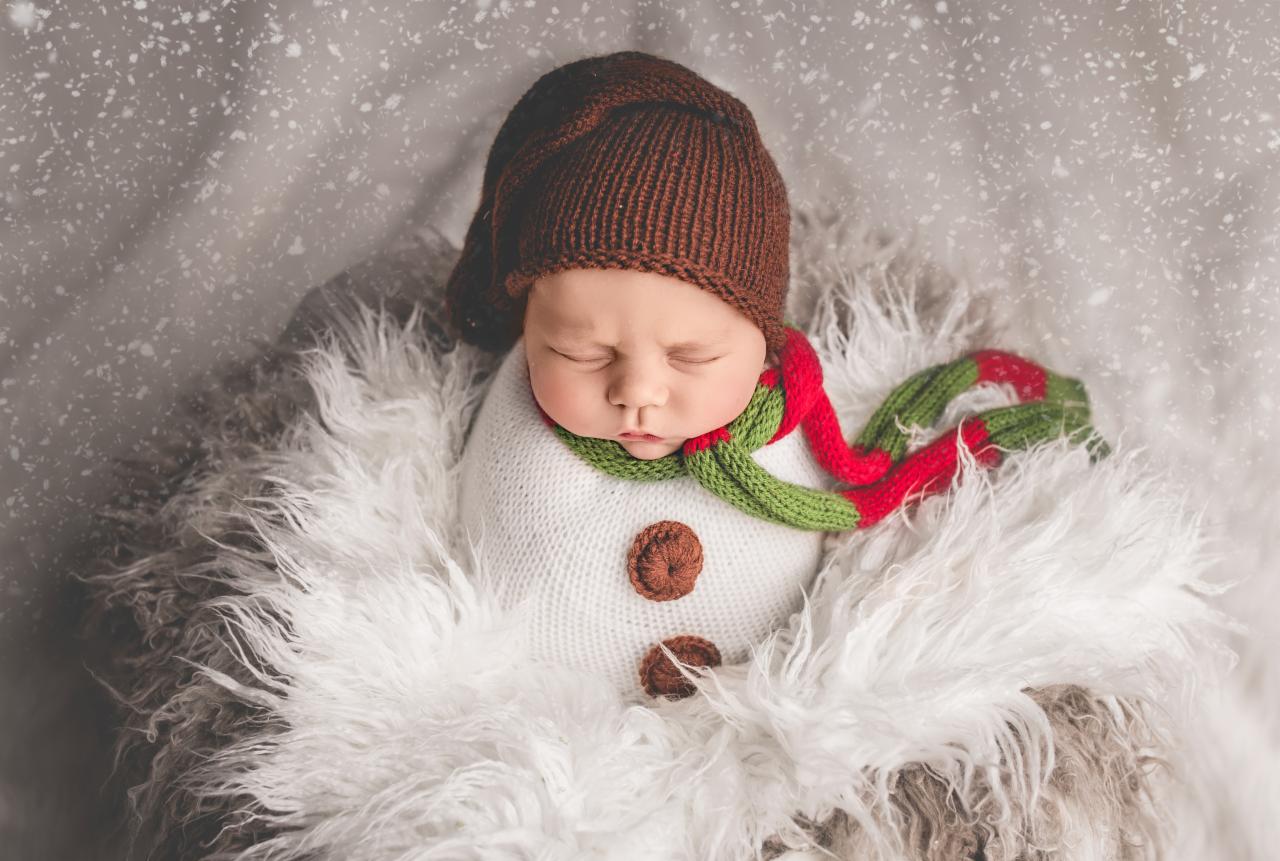 Śpiący noworodek w brązowej czapce, owinięty w biały materiał z guzikami, stylizowany na bałwana, z zielono-czerwonym szalikiem, na tle imitującym śnieg.