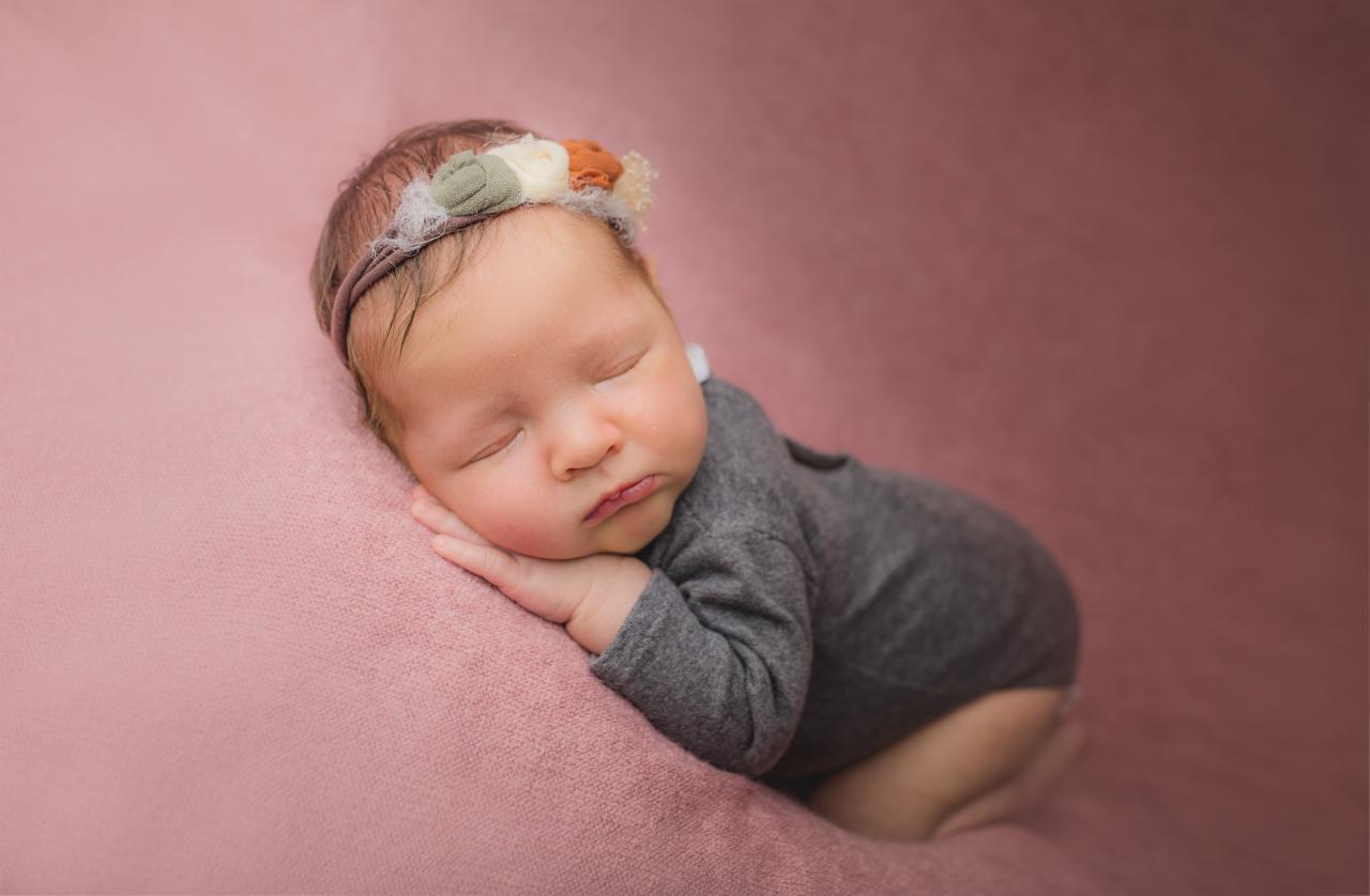 Śpiące niemowlę w szarym body i opasce z kwiatami na różowym tle.