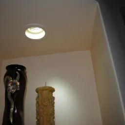 Wnęka z półką oświetloną halogenem, na której stoją dekoracyjne przedmioty: wysoki wazon z ornamentem i rzeźbiona świeca. Ściany w jasnym odcieniu beżu.