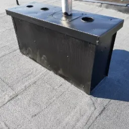 Szczelna obudowa komina z blachy na płaskim dachu pokrytym papą, z widocznymi otworami wentylacyjnymi i rurą kominową.