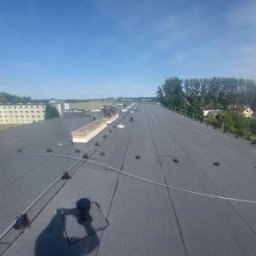 Łukasz Nowak - Rozległy, płaski dach pokryty papą termozgrzewalną z widocznymi elementami mocującymi i systemem odwadniającym. W tle budynki i zieleń.