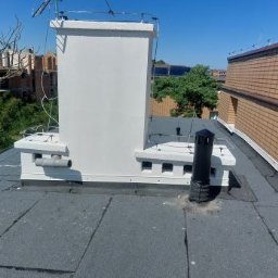 Dach pokryty papą z kominem i wentylacją. Widoczne elementy instalacji odgromowej. W tle budynki mieszkalne i błękitne niebo.