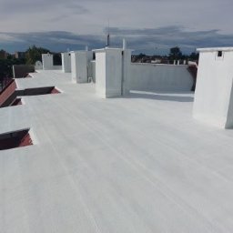 Biała, płaska powierzchnia dachu po renowacji z kominami wentylacyjnymi. Widok z góry, w tle zabudowa miejska i błękitne niebo z chmurami.