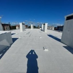 Biały, płaski dach budynku z kominami i wentylacją w słoneczny dzień. Widoczny cień osoby robiącej zdjęcie. Nowoczesne wykończenie dachu.