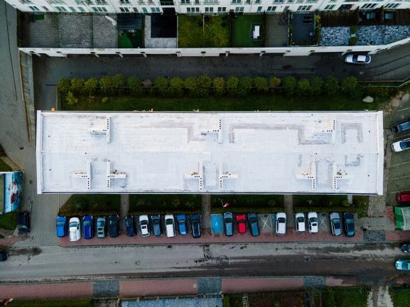 Widok z góry na płaski dach budynku pokryty białym materiałem izolacyjnym, z widocznymi elementami konstrukcyjnymi i parkingiem pełnym samochodów.