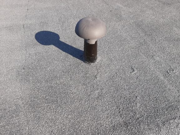 Szary odpowietrznik na pokryciu dachowym z papy, rzucający wyraźny cień w słoneczny dzień.