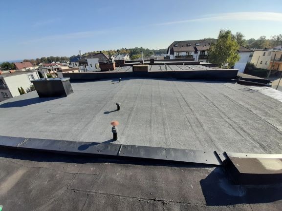 Płaski dach pokryty papą termozgrzewalną z widocznymi kominkami wentylacyjnymi i obróbkami blacharskimi, w tle zabudowa jednorodzinna i drzewa pod błękitnym niebem.