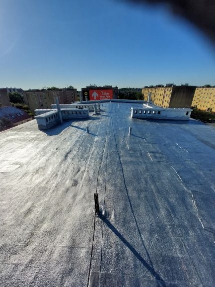 Płaski dach pokryty srebrzystą powłoką hydroizolacyjną, widoczne kominki wentylacyjne i budynki w tle pod błękitnym niebem.