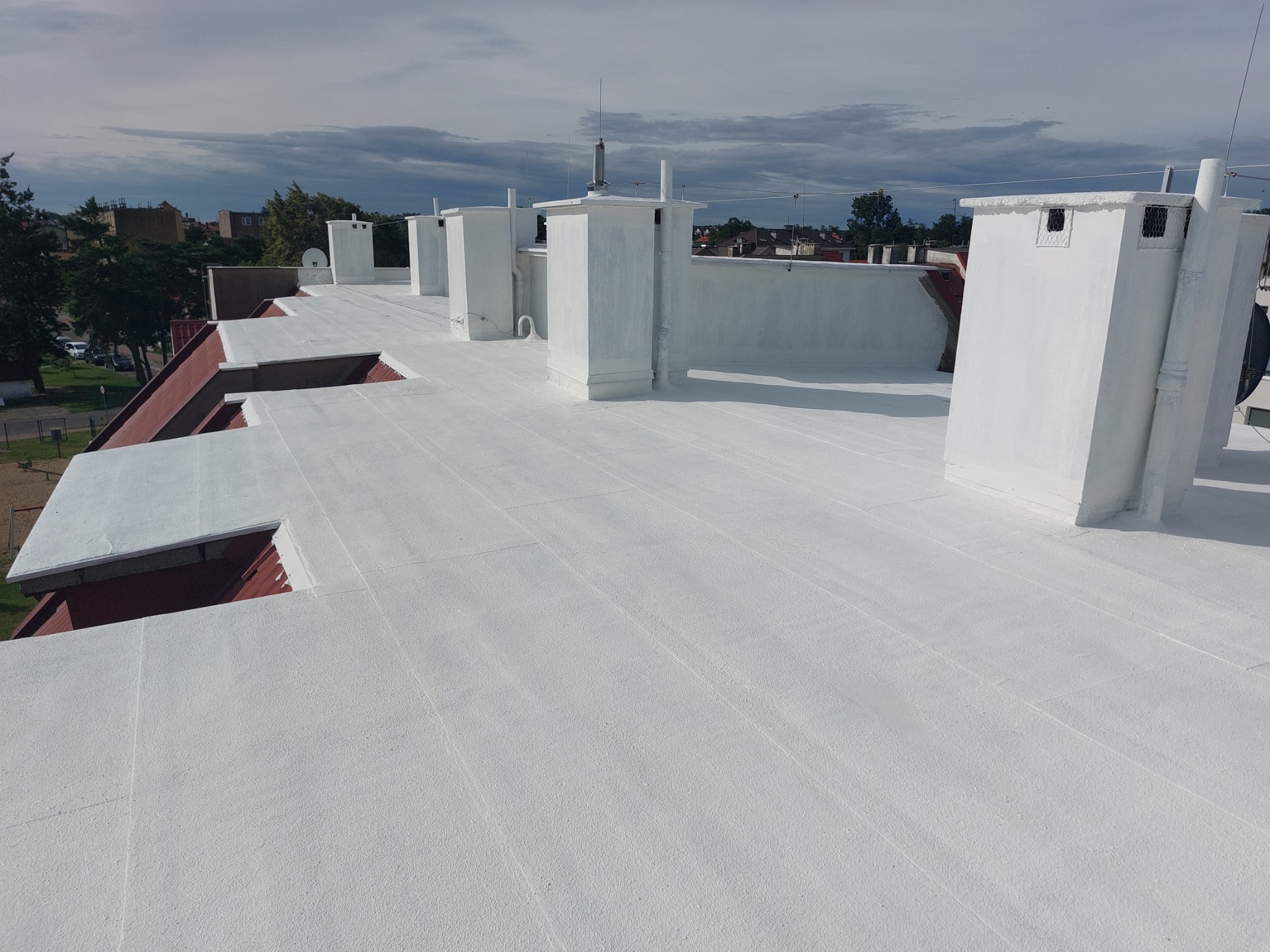 Biały dach po renowacji z kominami wentylacyjnymi na tle zabudowy miejskiej i pochmurnego nieba. Widok z góry.