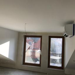 MarekProchal - Wykończone poddasze ze skośnym sufitem, widoczne dwa okna z brązowymi ramami zabezpieczone folią malarską, białe ściany z widocznymi śladami szpachlowania, zamontowany klimatyzator na ścianie...