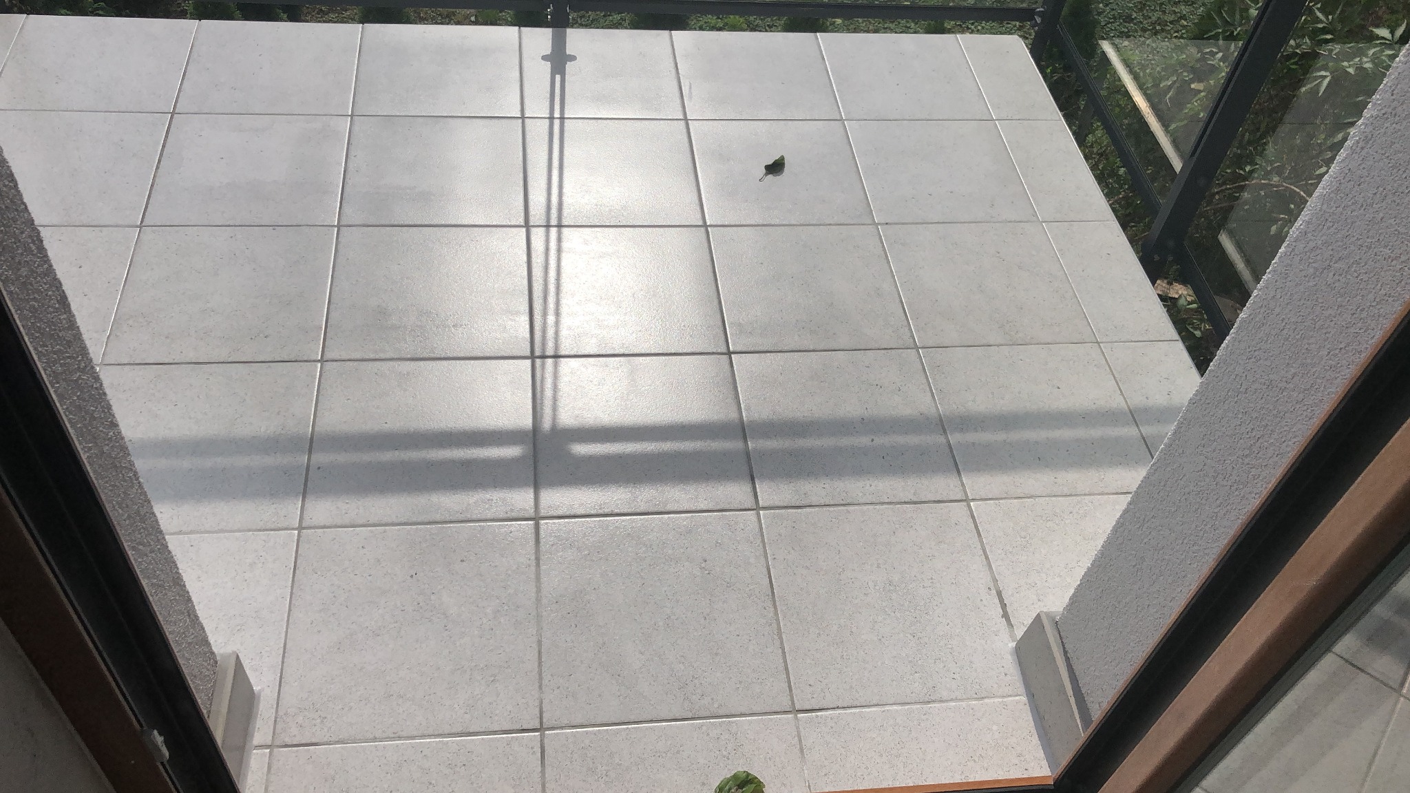 Jasnoszare płytki gresowe na balkonie, widok z góry przez otwarte drzwi balkonowe, cień barierki, pojedynczy liść na płytkach.