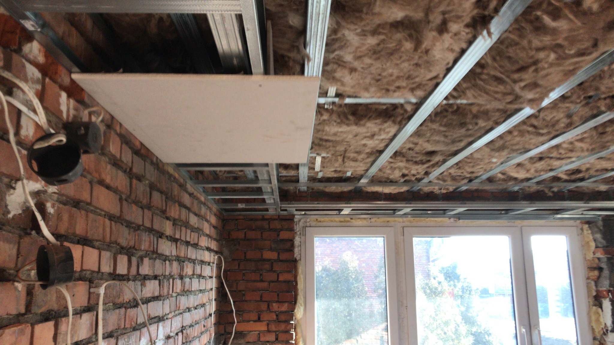 Montaż sufitu podwieszanego z płyt kartonowo-gipsowych na metalowej konstrukcji, widoczna izolacja z wełny mineralnej, ściana z czerwonej cegły z wystającymi przewodami elektrycznymi i puszki...