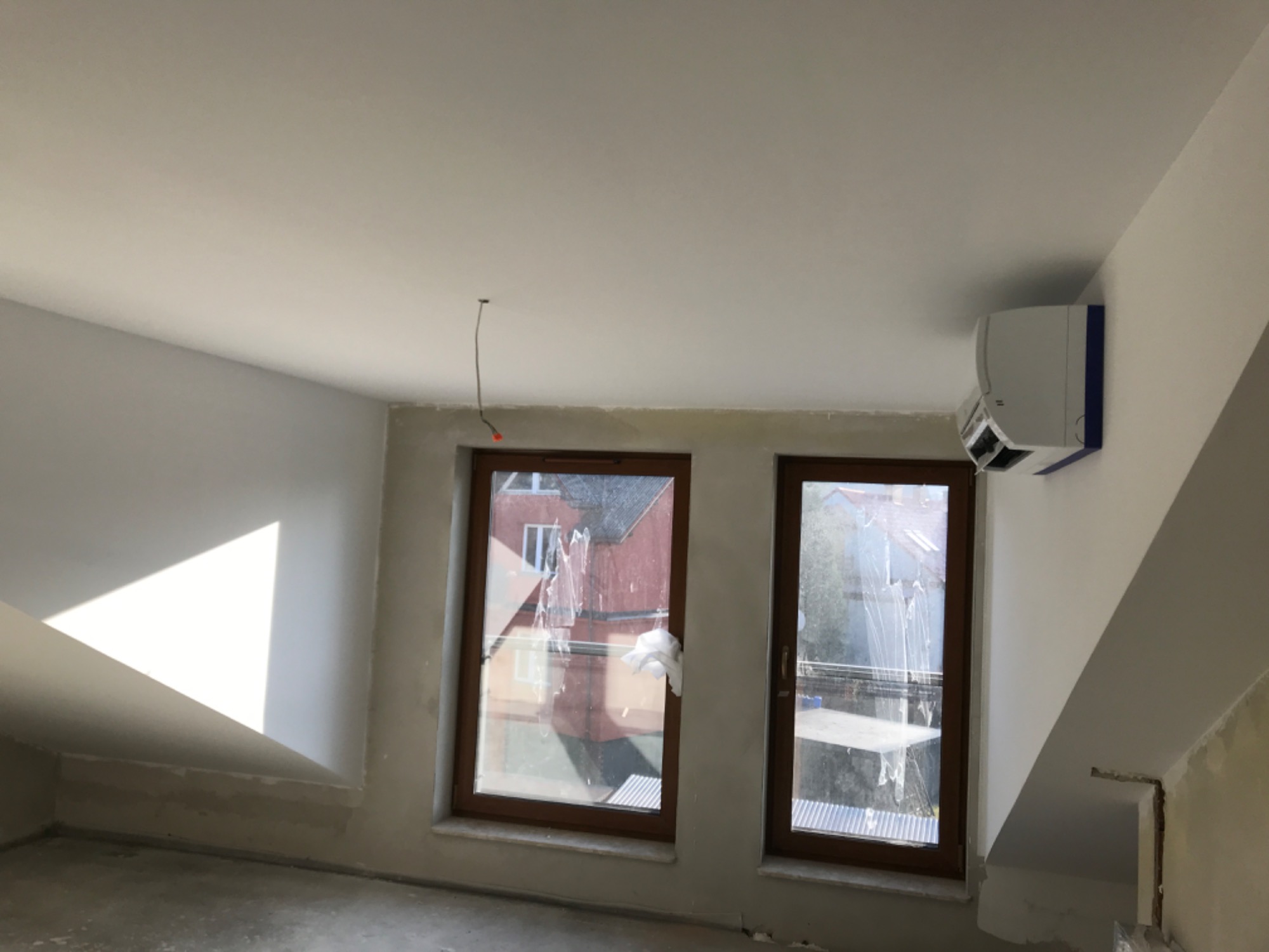 Wykończone poddasze ze skośnym sufitem, widoczne dwa okna z brązowymi ramami zabezpieczone folią malarską, białe ściany z widocznymi śladami szpachlowania, zamontowany klimatyzator na ścianie...