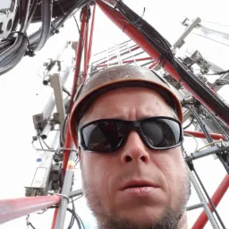 Mężczyzna w kasku i okularach na tle konstrukcji wieży telekomunikacyjnej. Widok z dołu, ukazujący kable i elementy montażowe.