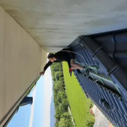 Monter instaluje rynnę na dachu pokrytym czarną dachówką, widok na zielone wzgórza w tle.