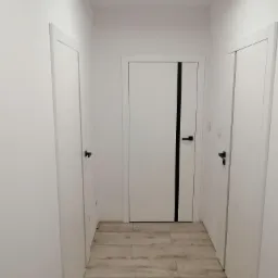 Korytarz z trzema białymi drzwiami z czarnymi klamkami, podłoga wyłożona panelami imitującymi drewno, minimalistyczny styl.