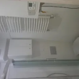 Biała łazienka z grzejnikiem drabinkowym, wiszącą szafką, toaletą i kabiną prysznicową z przezroczystym szkłem. Widoczny bojler nad grzejnikiem. Minimalistyczny styl.