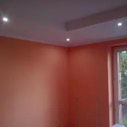 Pomarańczowa ściana w pokoju z białym sufitem podwieszanym z trzema punktowymi lampkami, widok przez okno na drzewo z jarzębiną.