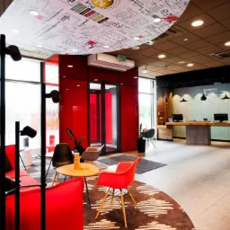 Wnętrze hotelu Ibis Poznań Północ z recepcją, strefą wypoczynkową z czerwonymi sofami i stolikami, oraz sufitem z grafikami i napisami.