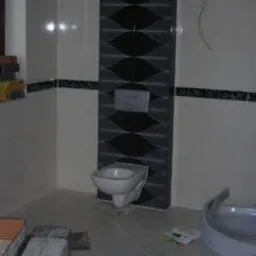 Wykończona łazienka w trakcie prac: białe płytki ścienne z czarnym pasem dekoracyjnym, fragment ściany z dekoracyjną mozaiką w odcieniach szarości i czerni, podwieszana toaleta, elementy wanny...