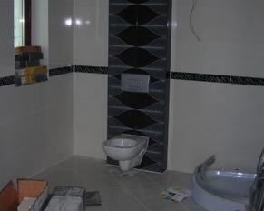 Wykończona łazienka w trakcie prac: białe płytki ścienne z czarnym pasem dekoracyjnym, fragment ściany z dekoracyjną mozaiką w odcieniach szarości i czerni, podwieszana toaleta, elementy wanny...